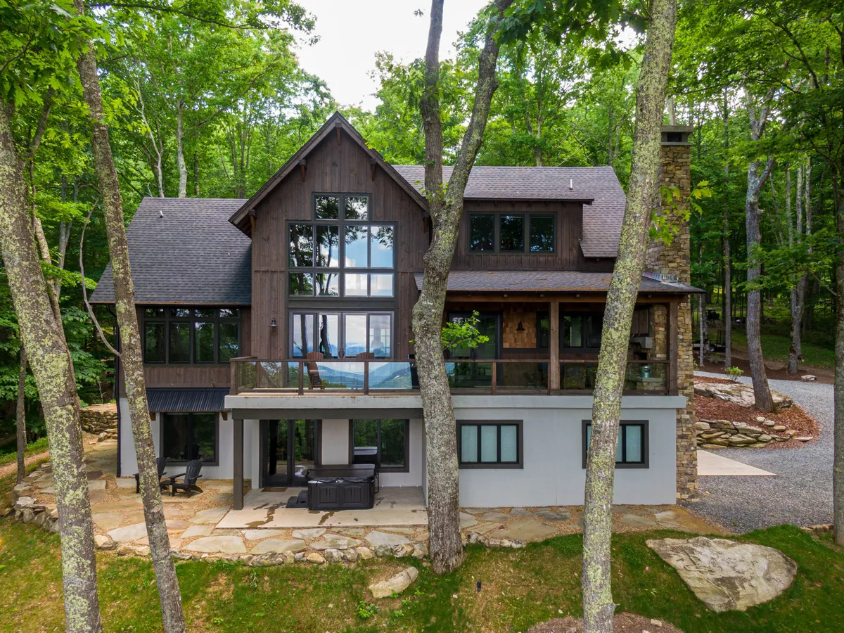 The hero image showcases Timber Forge Lodge’s striking wooden and stone exterior, framed by large glass windows that invite the outdoors in. The spacious outdoor seating area sits nestled among mature trees, offering mountain views and tranquil privacy—perfect for gathering with family, enjoying morning coffee, or relaxing by an evening fire.