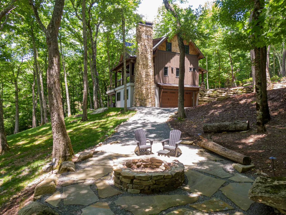 A serene fire pit area is featured here, with a stone fireplace, comfortable seating, and natural woods in the background. The arrangement of chairs invites guests to sit, socialize, and enjoy the ambiance of a crackling fire as dusk approaches.