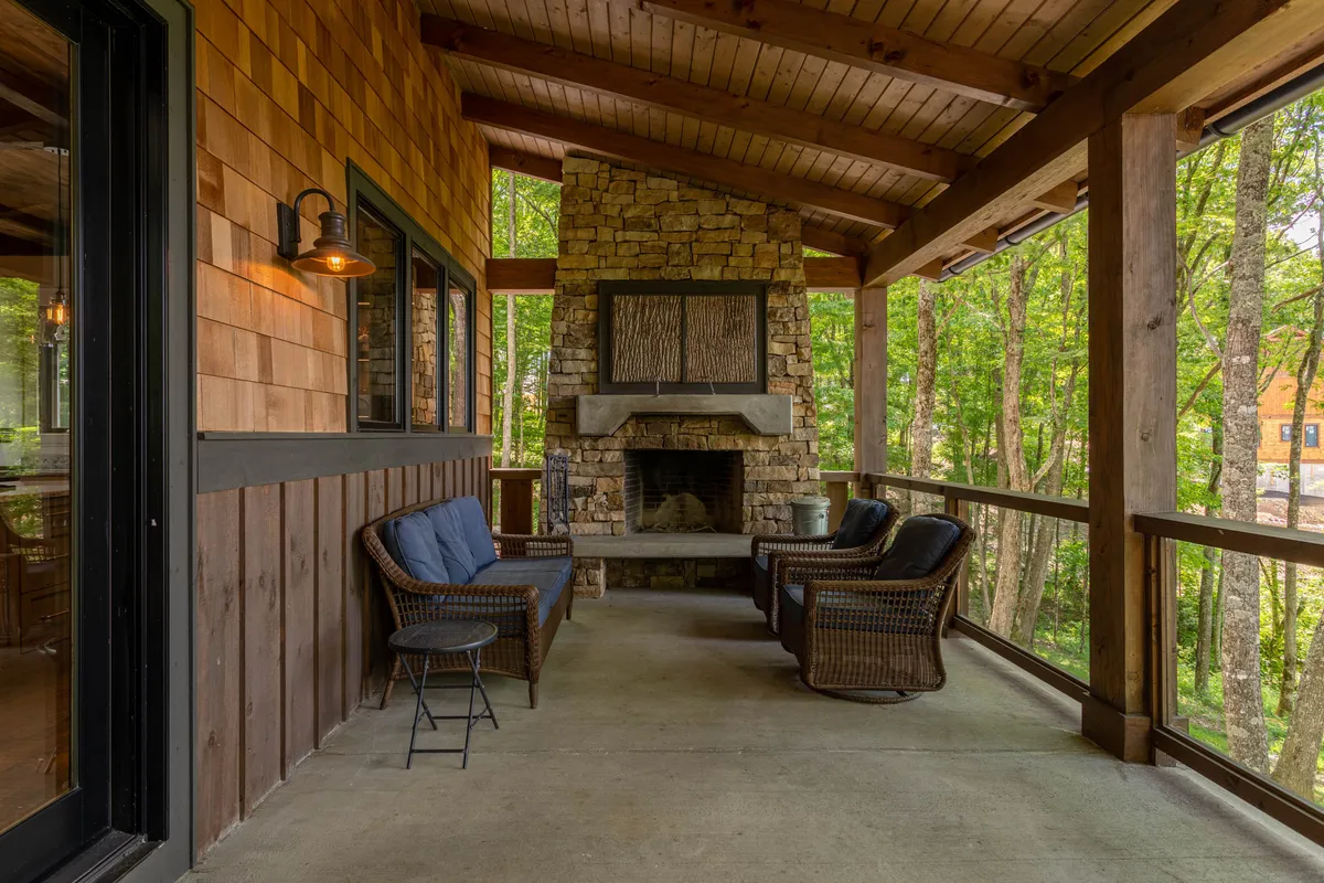 A stone outdoor fireplace anchors a covered deck surrounded by lush trees, with inviting wicker chairs arranged for group conversation or marshmallow roasting. The atmosphere is tranquil and cozy, with the green backdrop enhancing the sense of retreat and mountain escape.
