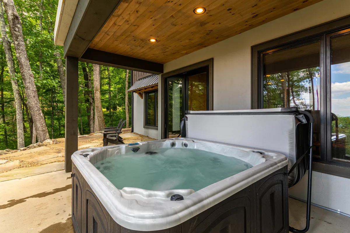 A tranquil deck nestled among tall trees, featuring a bubbling hot tub and comfortable seating. The deck is both private and inviting, offering a relaxing spot to unwind after an active day, with views of the surrounding forest.