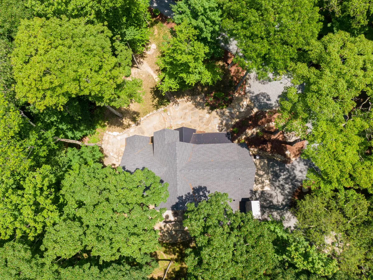 This image features the house surrounded by tall trees and a stone pathway leading into nature, suggesting a seamless flow between the home and the outdoors. The peaceful exterior environment hints at access to trails and the tranquility of the wooded setting.