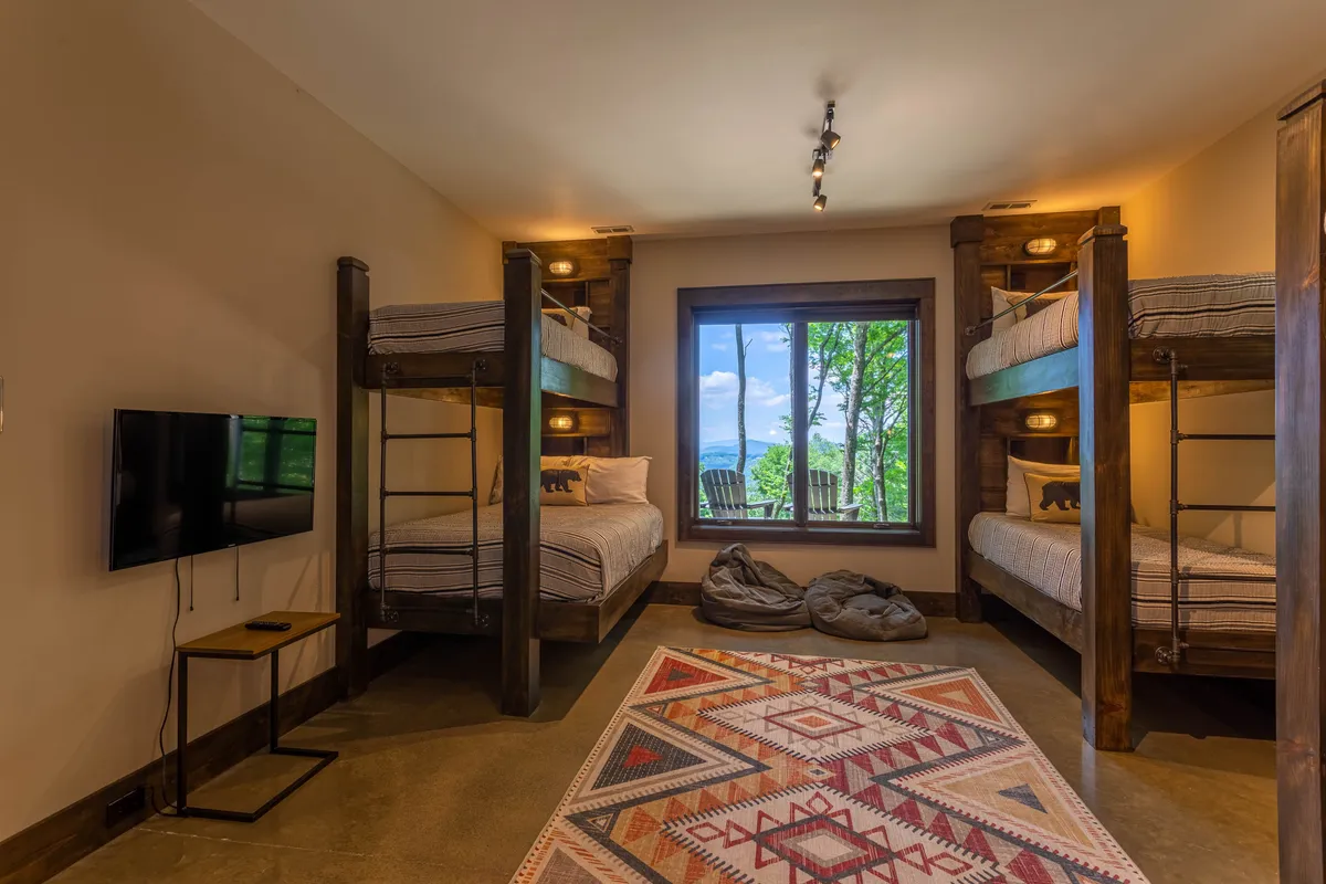 A playful bunk room featuring three sets of bunk beds, a large window that lets in natural light, a soft area rug, bean bags for lounging, and a television. The space is ideal for kids or singles, inviting both play and rest.