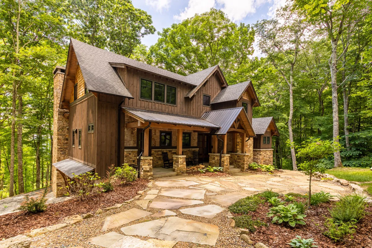 This exterior image shows a welcoming stone pathway leading up to a wooden porch with large windows overlooking a landscaped garden. The porch is arranged for relaxing or entertaining, and the scene is bathed in sunset light, emphasizing the lodge's inviting atmosphere and natural surroundings.