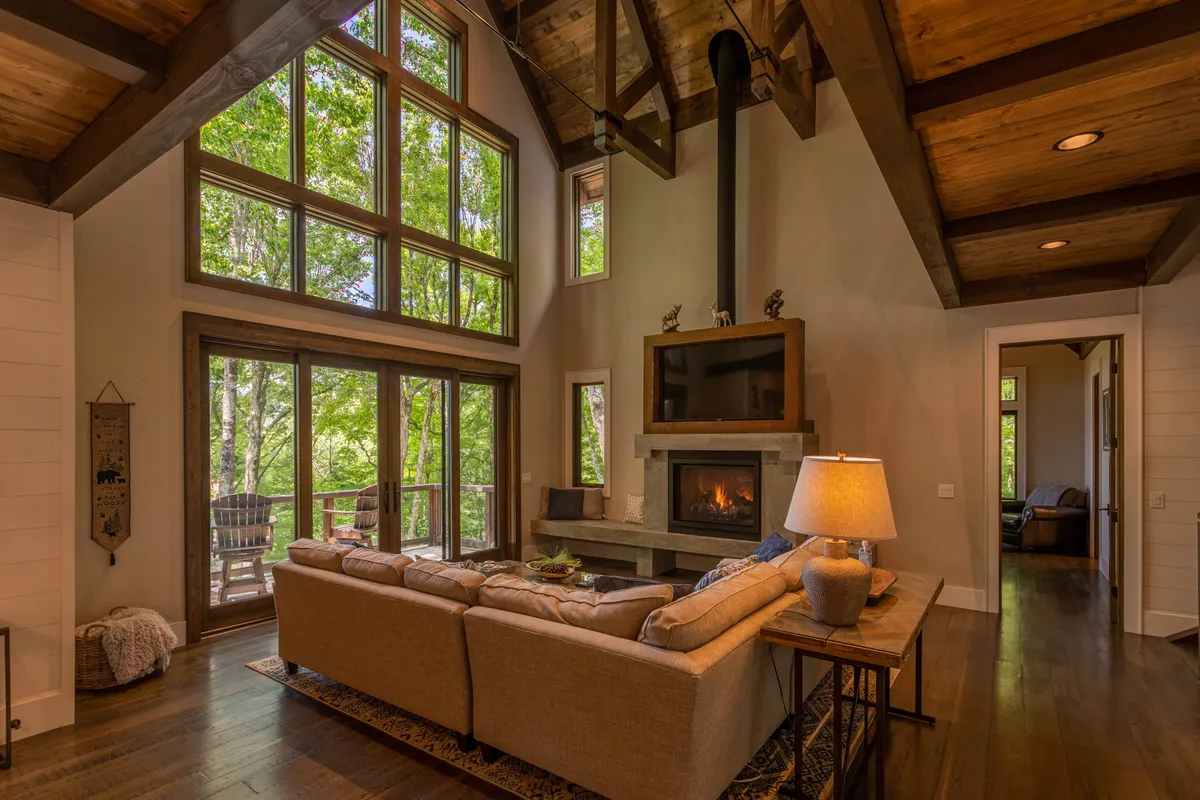 A cozy living room features a plush sectional sofa positioned to enjoy both a sleek modern fireplace and sweeping views through large windows. Wooden beams accent the ceiling, and the space exudes an atmosphere perfect for relaxing, reading, or gathering for conversation or a movie night.