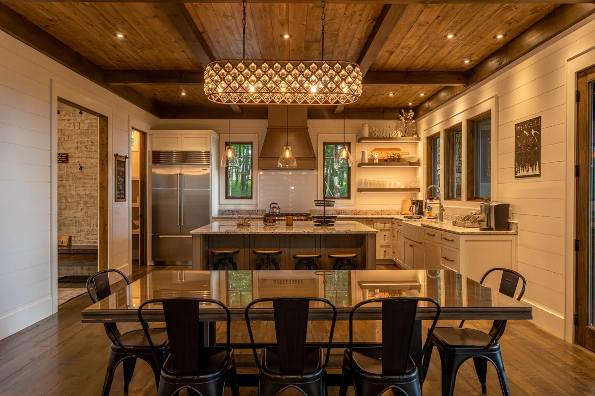 An open-concept dining and kitchen area features a large central table that seats a group, illuminated by an elegant pendant light. The kitchen island with bar stools and open shelving create a communal, celebratory atmosphere that hints at both intimate dinners and larger gatherings. Decorative items and thoughtful design touches emphasize comfort and togetherness.