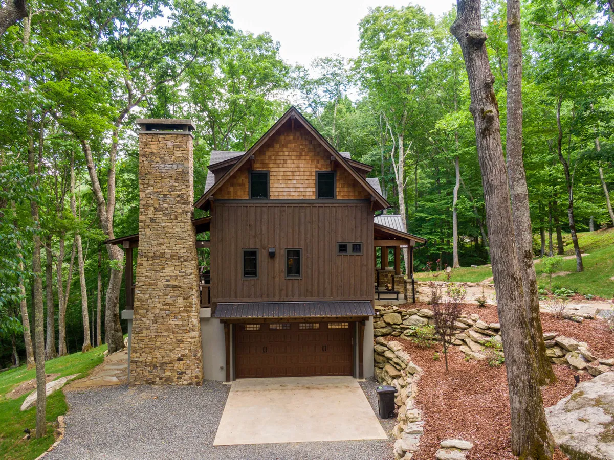 A cozy exterior shot of Timber Forge Lodge featuring a natural stone chimney, warm wooden siding, and surrounded by mature trees. The gravel driveway and the inviting porch create a welcoming transition between the home and the outdoors.