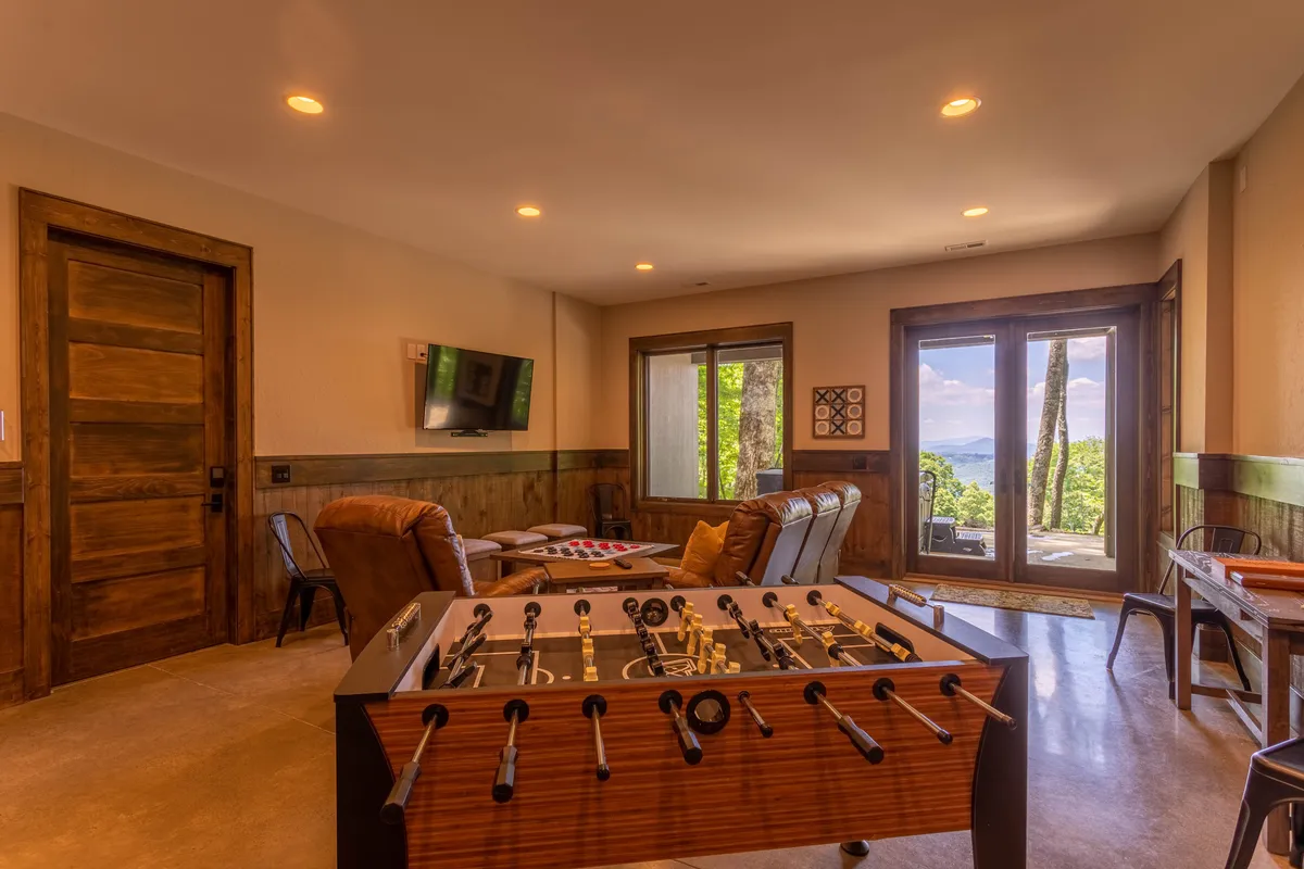 A recreational lounge area with a foosball table, comfortable seating, large windows offering an outdoor view, and a TV mounted on the wall, providing a playful yet cozy environment ideal for both games and relaxation.