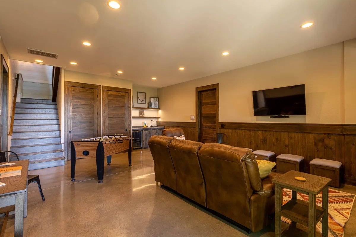 A cozy downstairs family room featuring a brown leather sofa, foosball table, staircase leading upstairs, a television mounted on the wall, and wooden cabinets, all arranged in an open, welcoming setting suitable for socializing and entertainment.